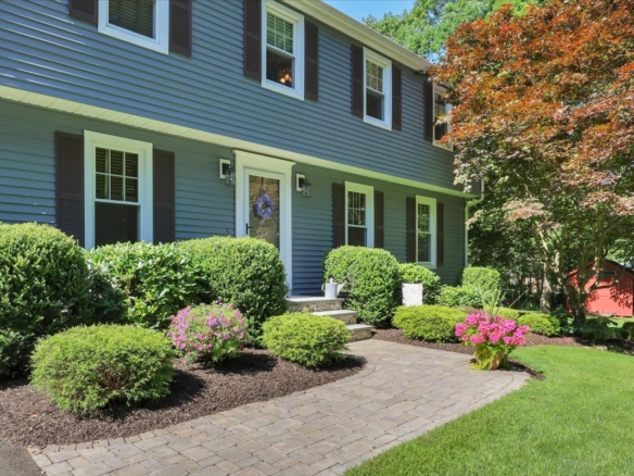 A close-up of a paved stone walkway and professional landscaping leading to the front door of a suburban home, featuring flowering plants and decorative details.