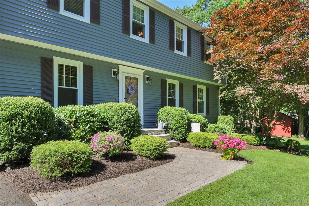 A close-up of a paved stone walkway and professional landscaping leading to the front door of a suburban home, featuring flowering plants and decorative details.