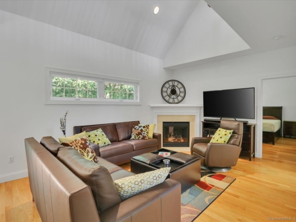 Bright family room with a cathedral ceiling, brown leather conversational seating, a glass-door corner fireplace, a large flat-screen TV, and an adjoining bedroom entrance.