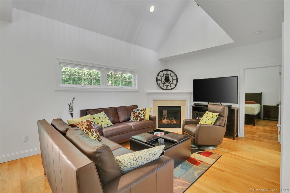 Bright family room with a cathedral ceiling, brown leather conversational seating, a glass-door corner fireplace, a large flat-screen TV, and an adjoining bedroom entrance.