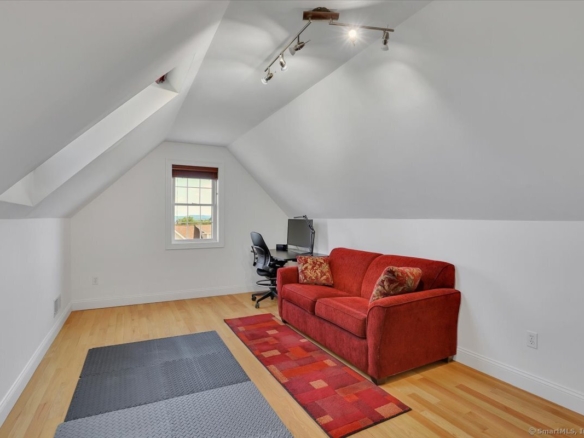 Finished attic bonus room with sloped ceilings, a cozy red plush sofa, a desk station, and a dormer window showcasing a distant view.