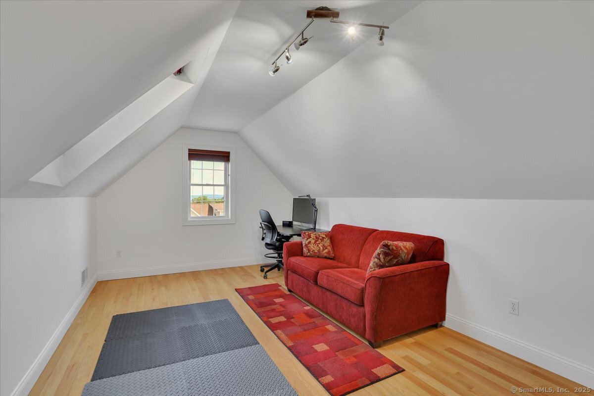 Finished attic bonus room with sloped ceilings, a cozy red plush sofa, a desk station, and a dormer window showcasing a distant view.