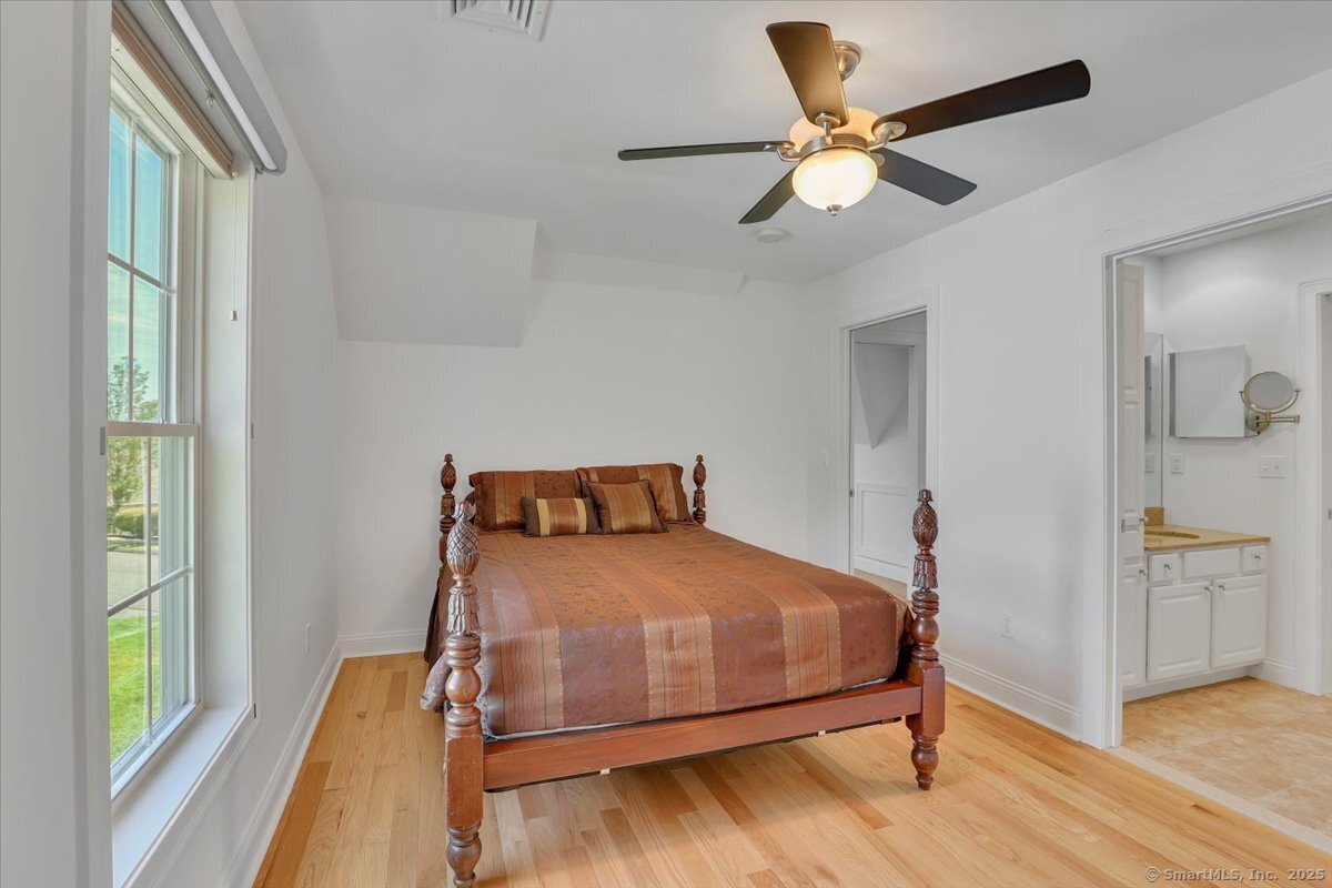 Bright second bedroom with an ornate four-poster wooden bed, natural wood flooring, a ceiling fan, and an attached white vanity bathroom entrance.