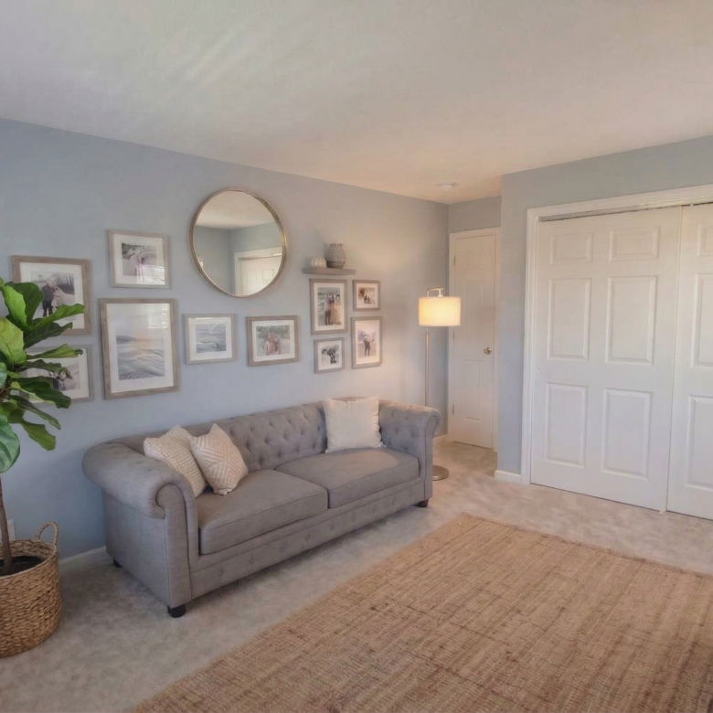 A virtually staged den featuring blue-grey walls, a grey tufted sofa, a large fiddle leaf fig plant, and a gallery wall with a round mirror.
