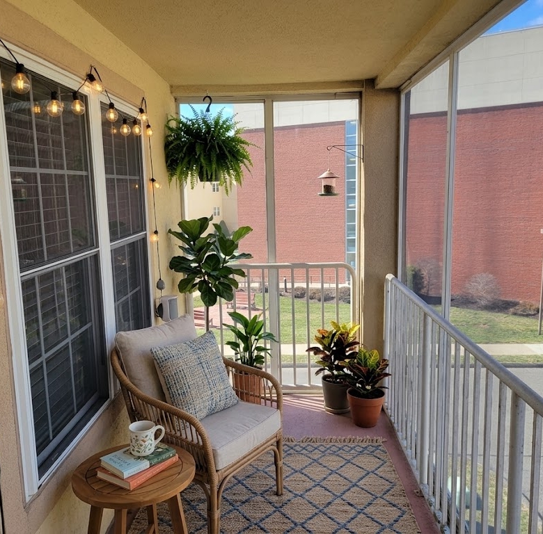 A cozy screened-in balcony staged with a wicker chair, string lights, outdoor rug, and various potted plants.