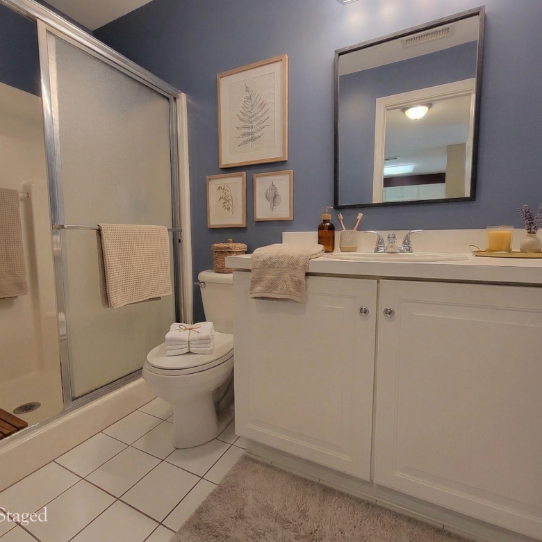 A bathroom with deep blue walls, a white vanity, a walk-in shower with sliding glass doors, and virtually staged decor.