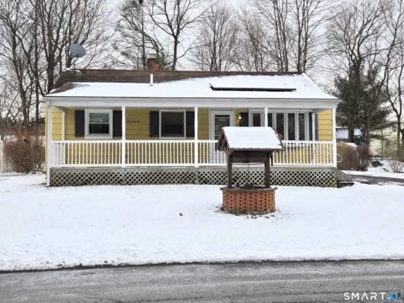 Front exterior of a yellow ranch-style house with a full-length white porch and a decorative brick wishing well in the snow.