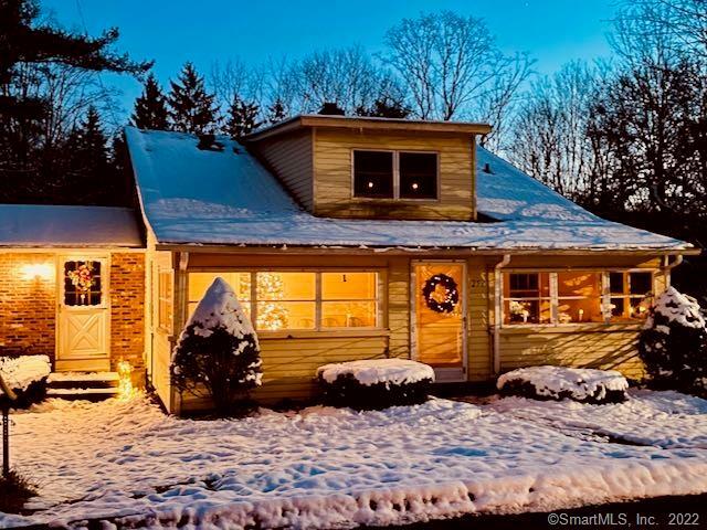 A cozy, light-yellow cottage-style home adorned with festive holiday decorations, glowing warm interior lights, and a light dusting of snow during the winter season in Durham, CT.