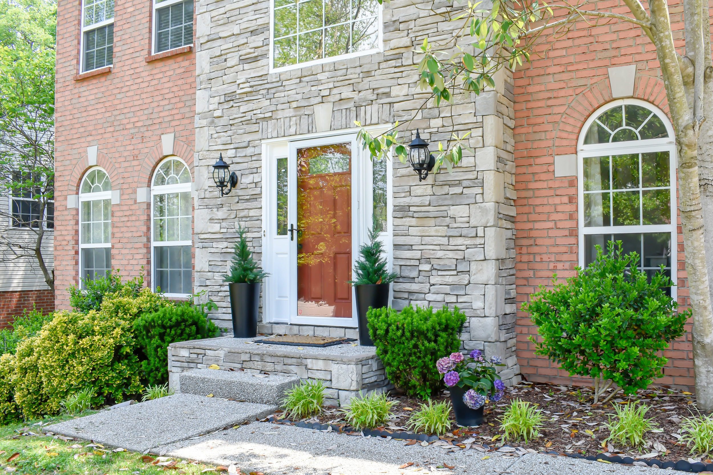 Front entry of a custom brick and stone colonial home in Wallingford, Connecticut, featuring arched windows, a red front door, and professionally landscaped flower beds.