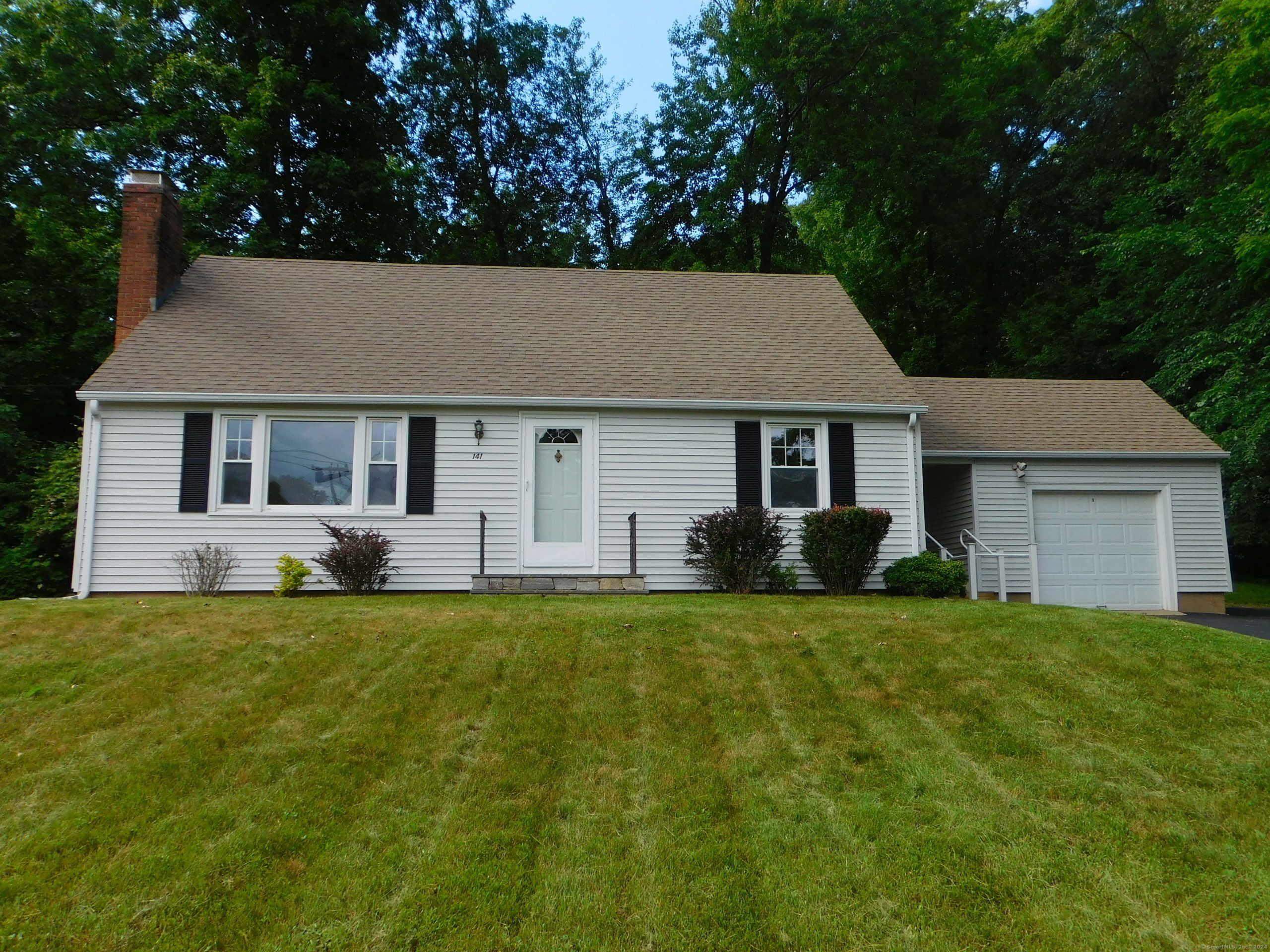 Exterior view of a charming white Cape Cod style home featuring black shutters, a brick chimney, a two-car garage, and a peaceful, tree-lined setting.