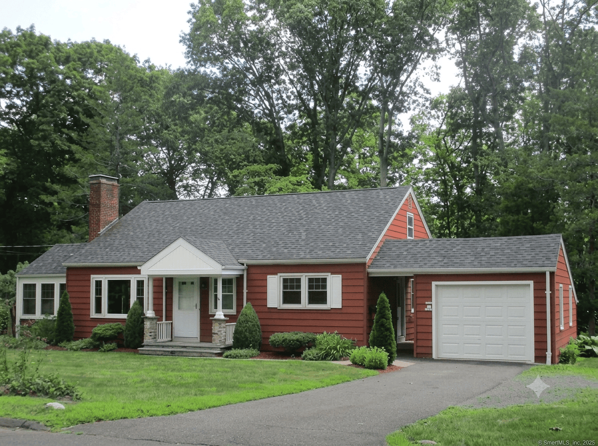 A charming red Cape Cod style home for sale in Wallingford, CT, featuring white trim, a covered front porch, and an attached garage set against a lush, wooded background.