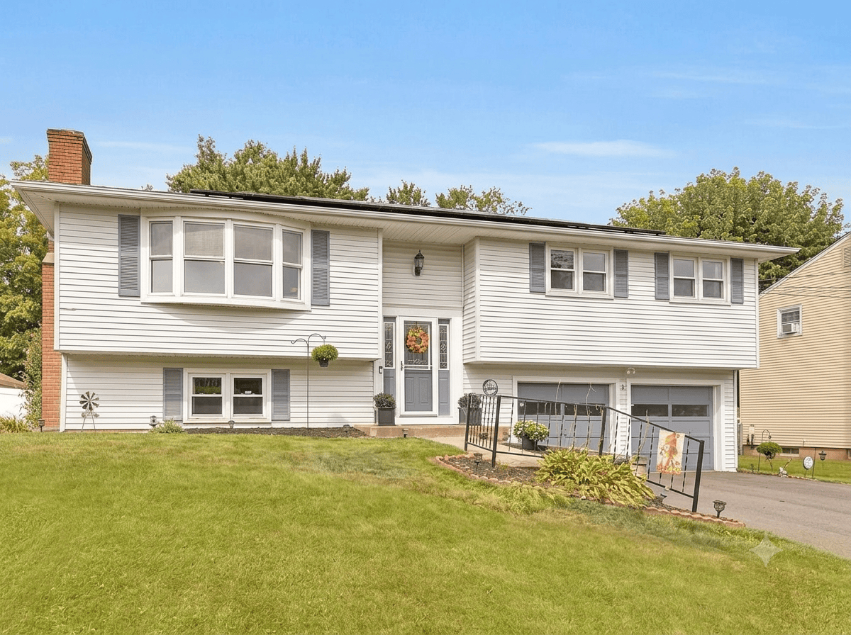 Exterior view of a well-maintained, classic split-level (split-foyer) home in Meriden, Connecticut, with white siding, light-blue shutters, a two-car garage, and a large bay window, set under a clear blue sky.