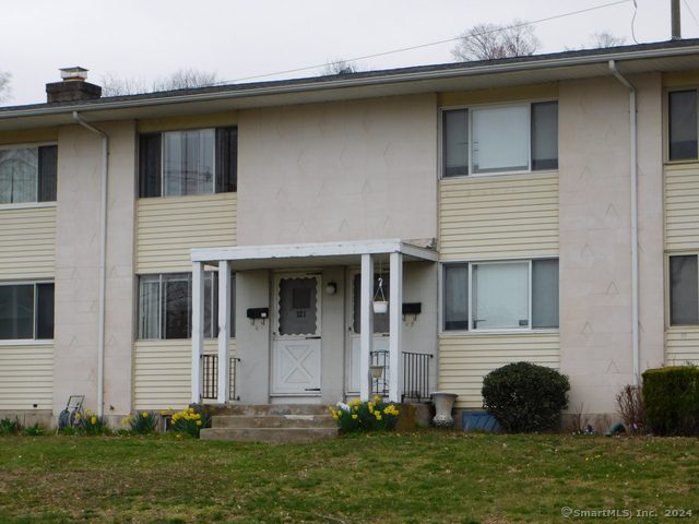 Exterior view of a condo for sale in Wallingford, Connecticut, featuring neutral siding, a covered entryway, and landscaped front lawn with spring flowers.