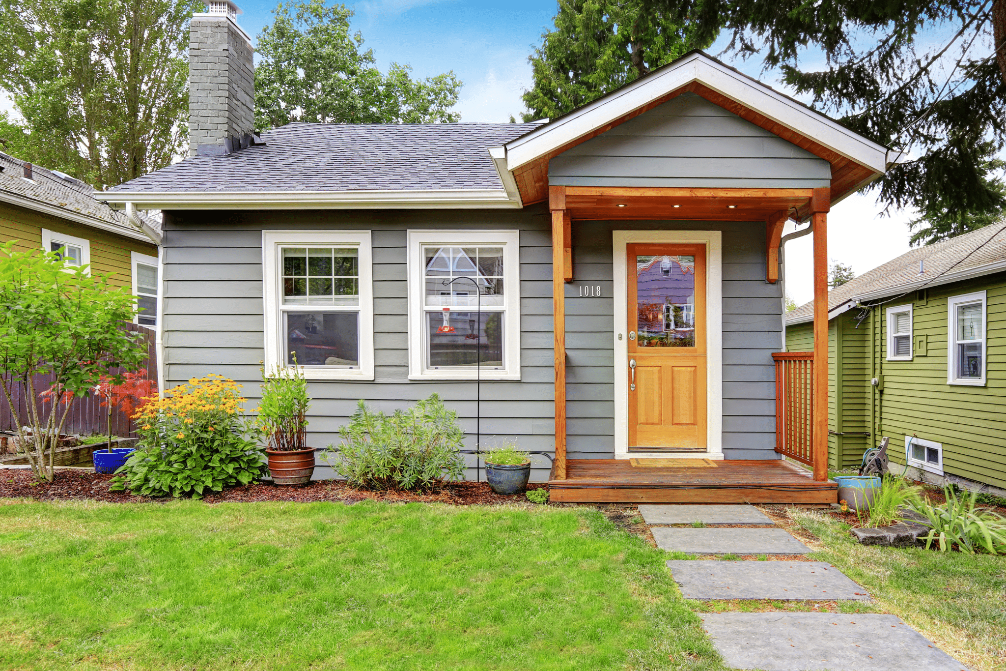 cozy, grey-sided bungalow featuring a warm natural wood front porch, a flagstone walkway, and vibrant, low-maintenance garden beds.