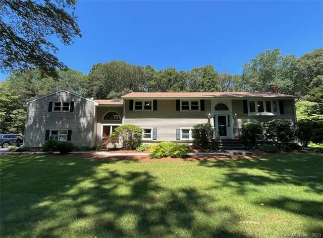 A spacious colonial-style home for sale in Cheshire, CT, featuring light siding, dark shutters, a large front lawn, and a mature tree-lined backyard under a clear blue sky.