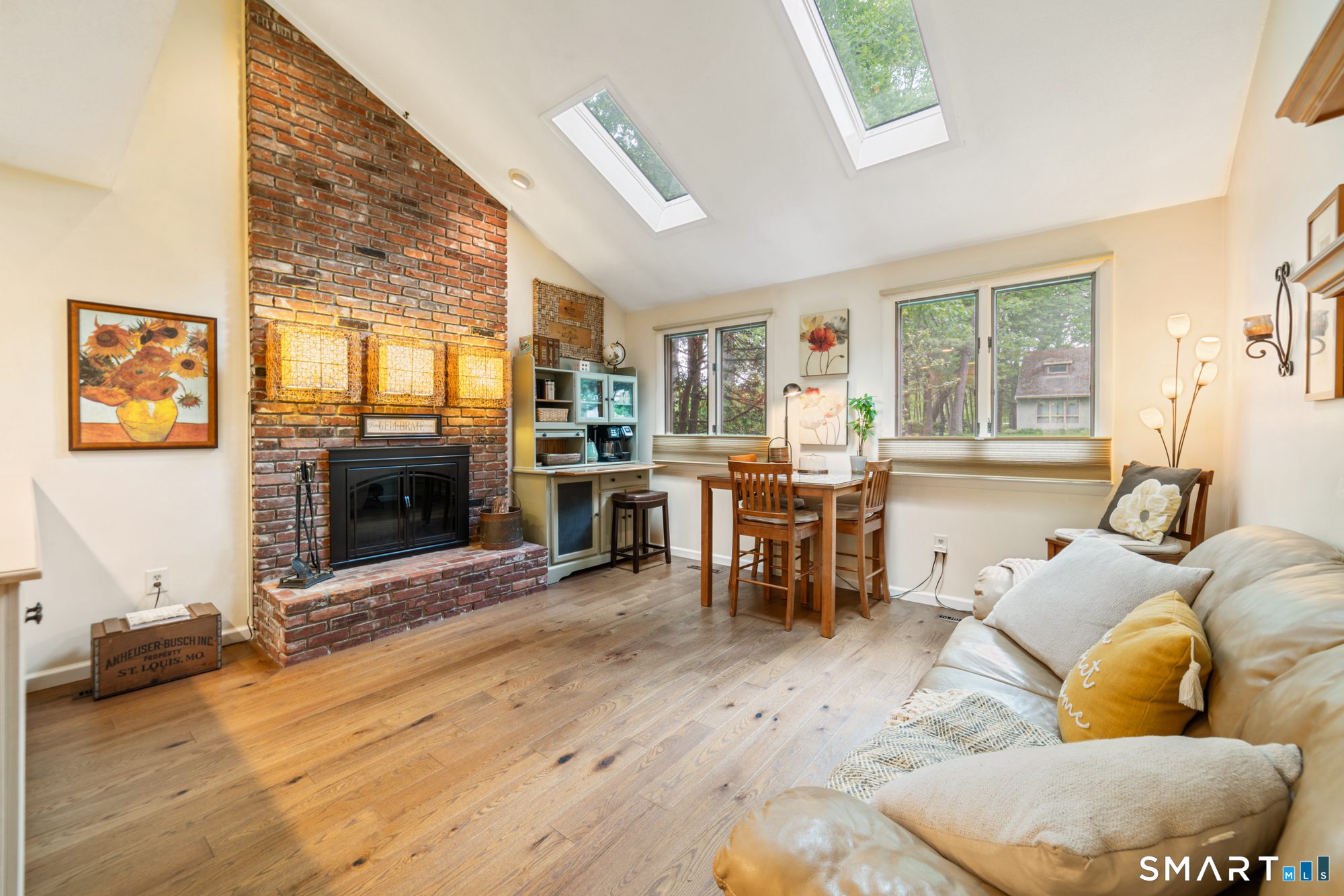 Sun-filled den featuring skylights, a brick fireplace, hardwood floors, and a small dining nook.